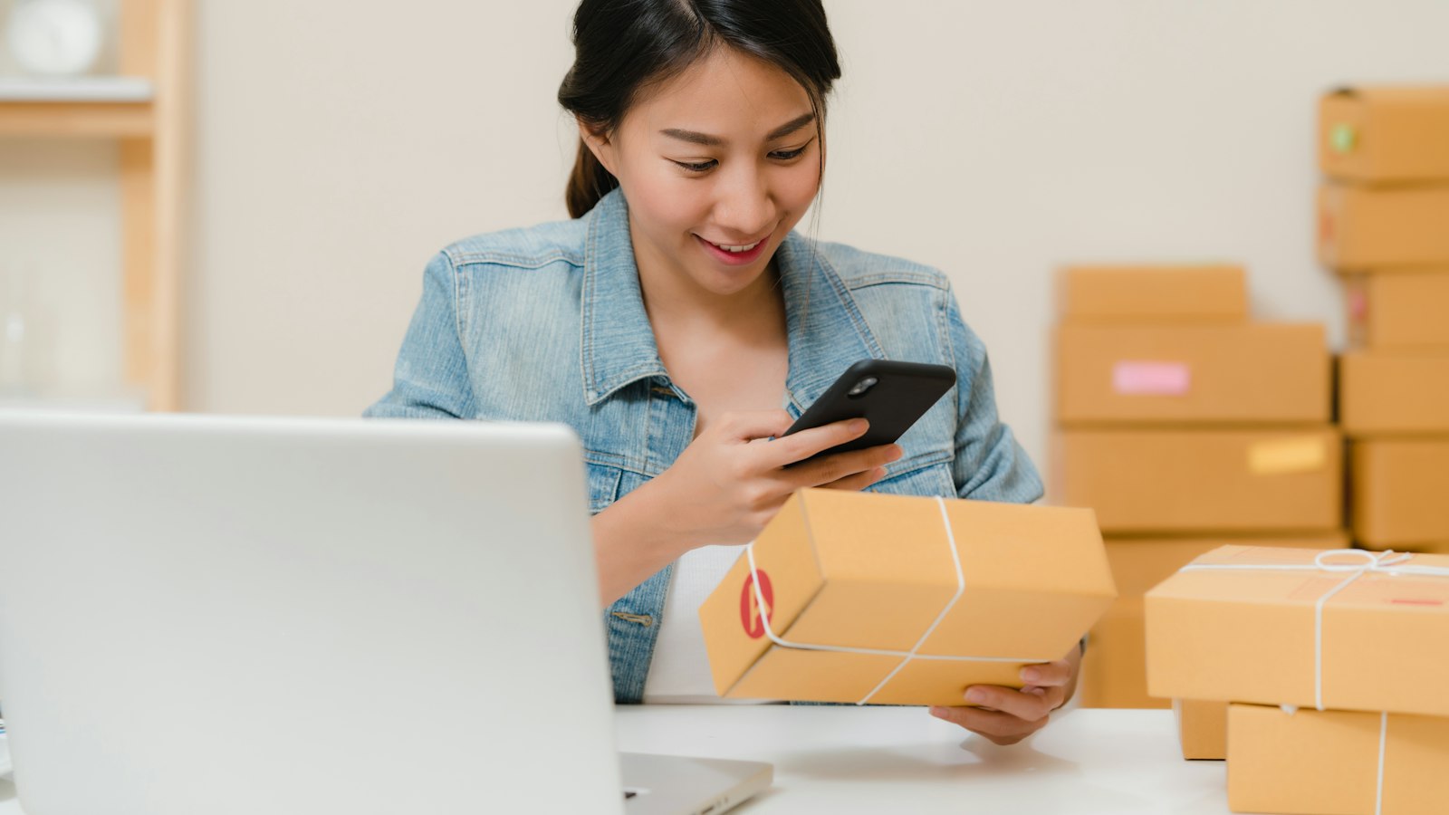 Une vendeuse en ligne consulte son téléphone en préparant des colis à côté de son ordinateur portable