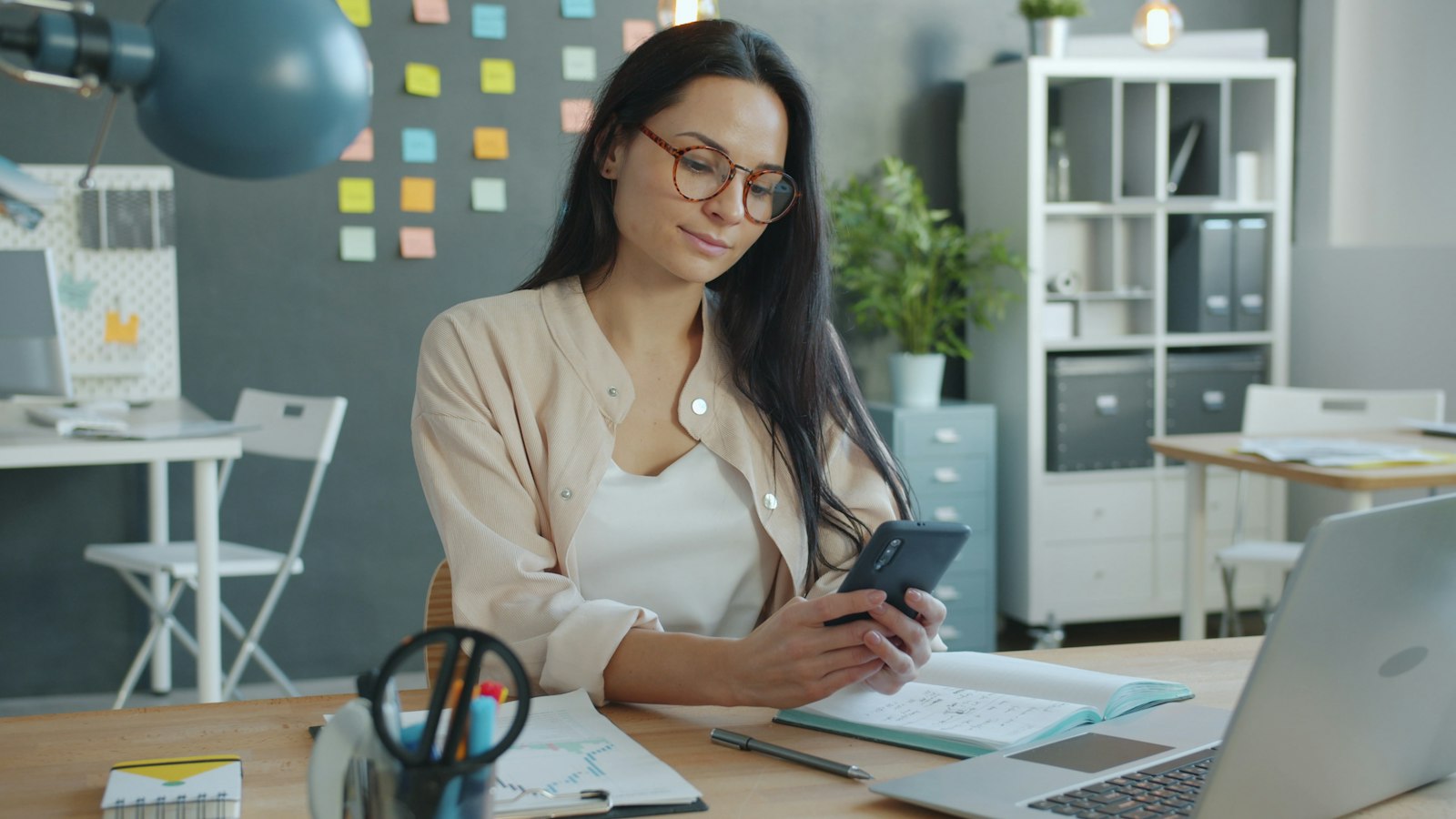 Une femme consulte les statistiques de sa page sur son téléphone, assise à son bureau devant un ordinateur portable